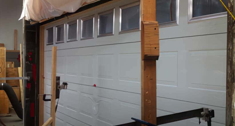 A white garage door with rectangular windows at the top is partially open and supported by wooden beams and clamps inside a workshop, suggesting ongoing garage door repair. Tools and equipment are visible around the area.