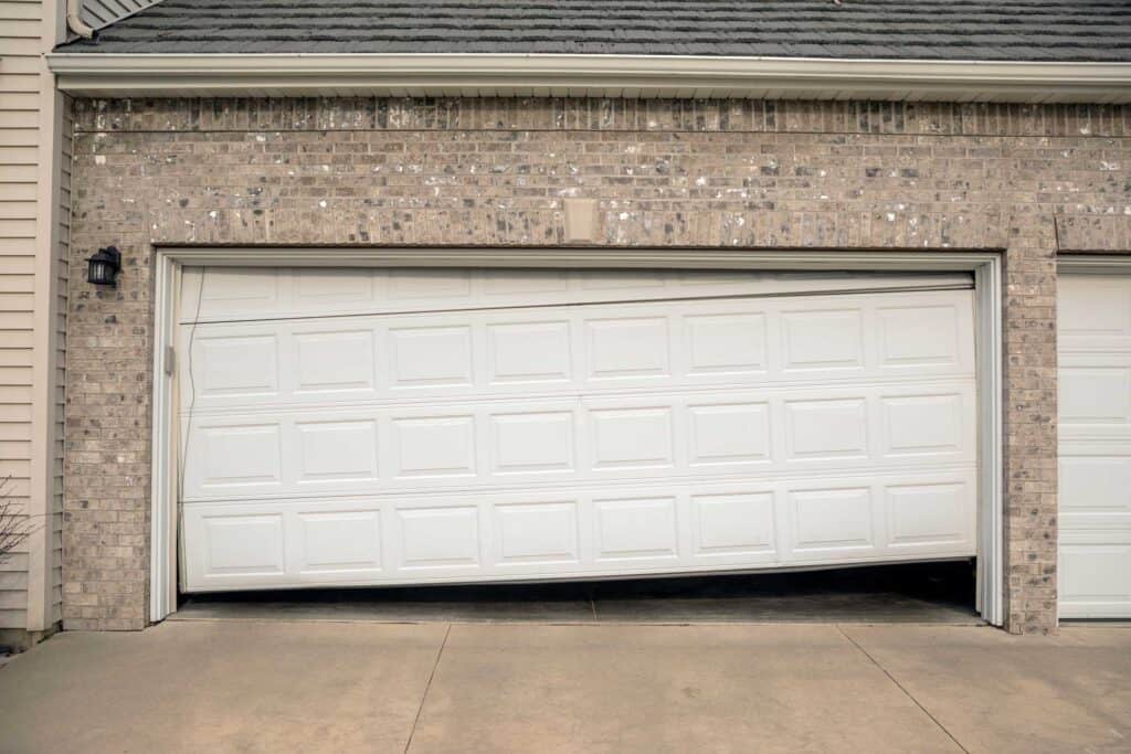 A white garage door is partially open and crooked, with one side higher than the other, appearing broken or off track. The garage doors are part of a brick house with a concrete driveway, possibly needing attention from a garage door company.