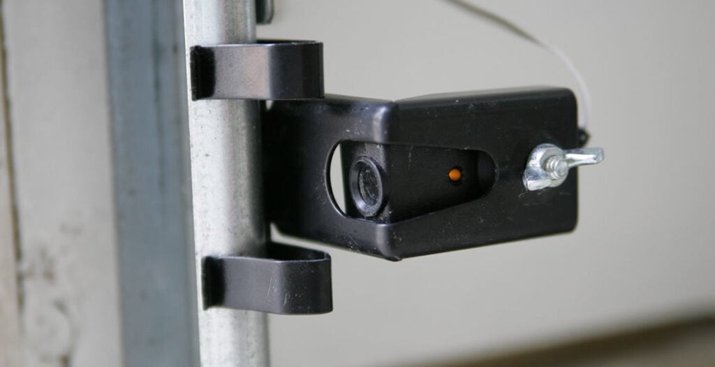 Close-up of a garage door safety sensor attached to a metal rail, featuring a small black housing with a lens and an orange indicator light—vital for the garage door auto reverse feature.