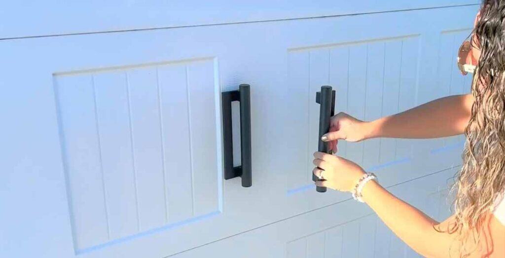 A person with wavy hair and a beaded bracelet is applying a magnetic garage door hardware black handle on a white garage door.